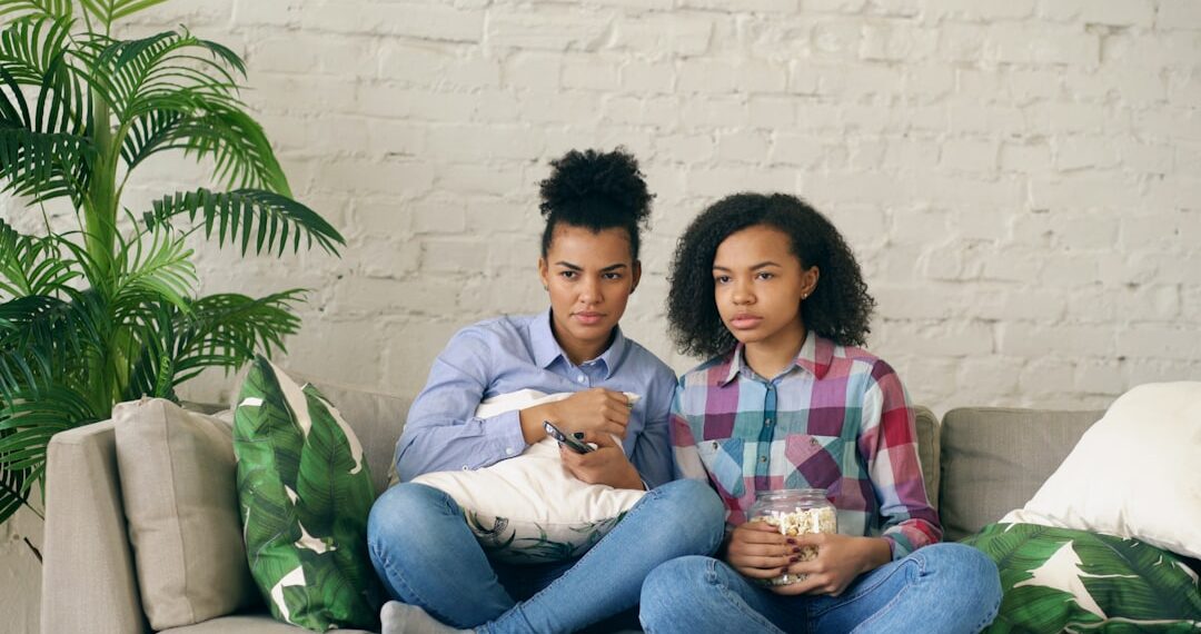 Two mixed race curly girl friends sitting on the couch and watch very scary movie on TV and eat popcorn