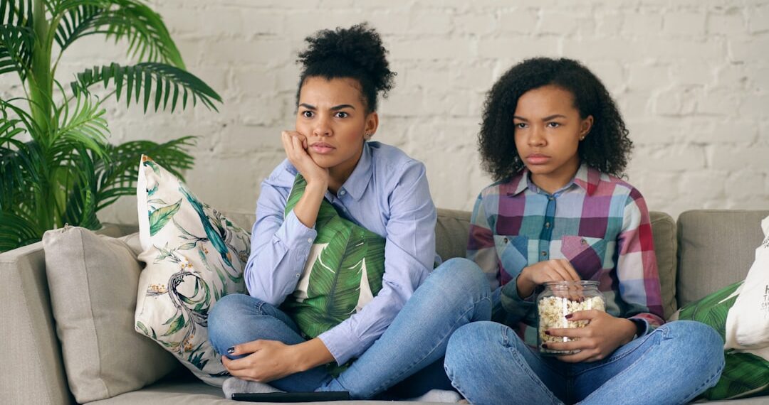 Two mixed race curly girl friends sitting on the couch and watch thriller movie and eat popcorn