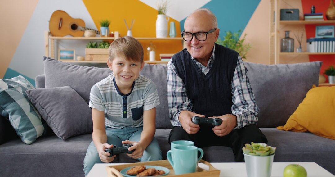 Grandfather and grandson are playing video game sitting on sofa at home using joysticks enjoying entertainment. Family and modern technology concept.