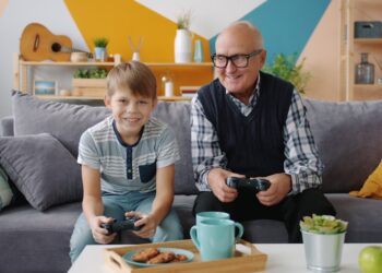 Grandfather and grandson are playing video game sitting on sofa at home using joysticks enjoying entertainment. Family and modern technology concept.