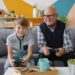 Grandfather and grandson are playing video game sitting on sofa at home using joysticks enjoying entertainment. Family and modern technology concept.