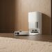A sleek white robotic vacuum cleaner is parked at its self-emptying and charging station in a modern, minimalist room with light hardwood floors and simple decor.
