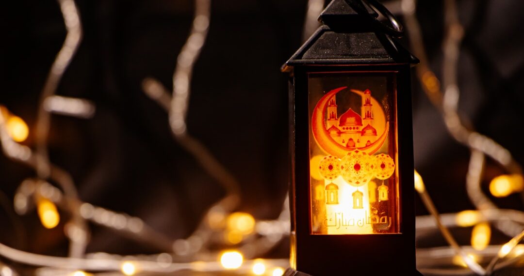 A lit black Ramadan lantern with a mosque design stands against a background of warm bokeh fairy lights.