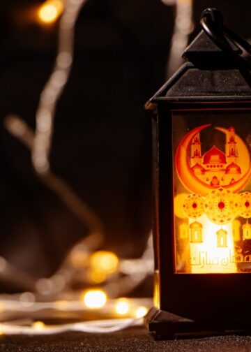 A lit black Ramadan lantern with a mosque design stands against a background of warm bokeh fairy lights.