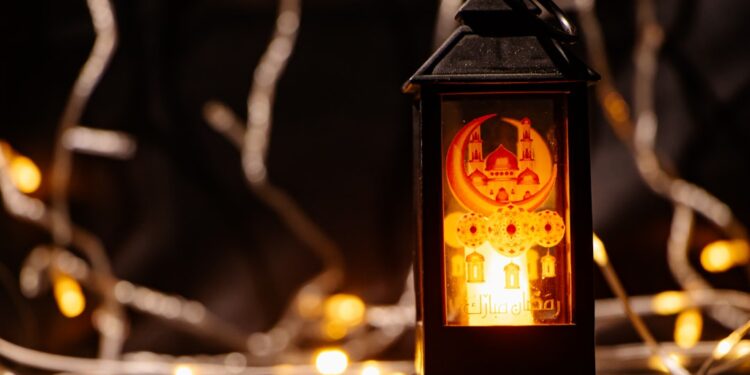 A lit black Ramadan lantern with a mosque design stands against a background of warm bokeh fairy lights.