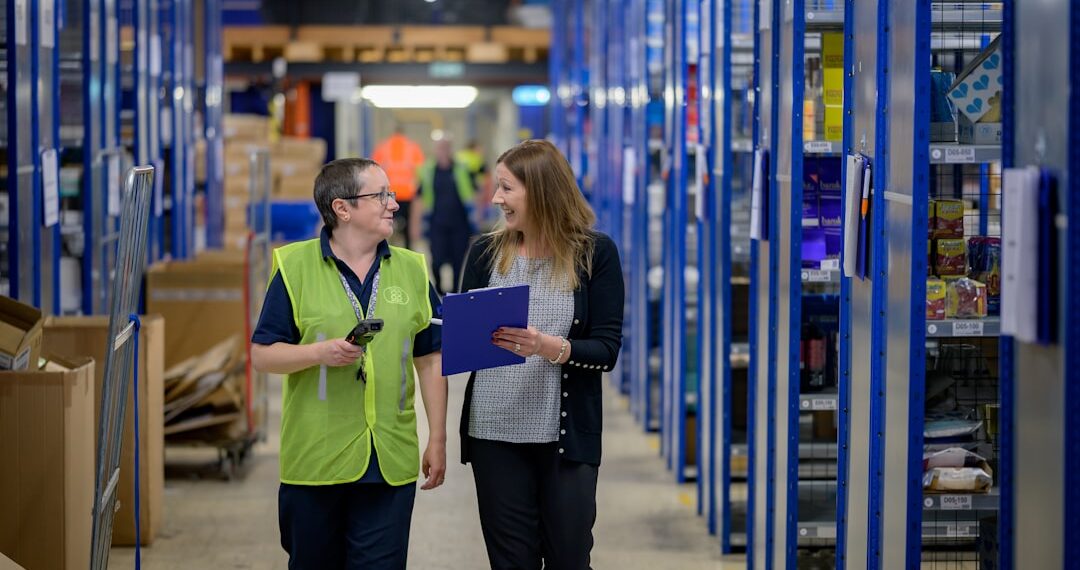 Two women walking through a warehouse and talking. One is holding a clipboard.