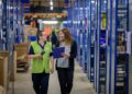 Two women walking through a warehouse and talking. One is holding a clipboard.