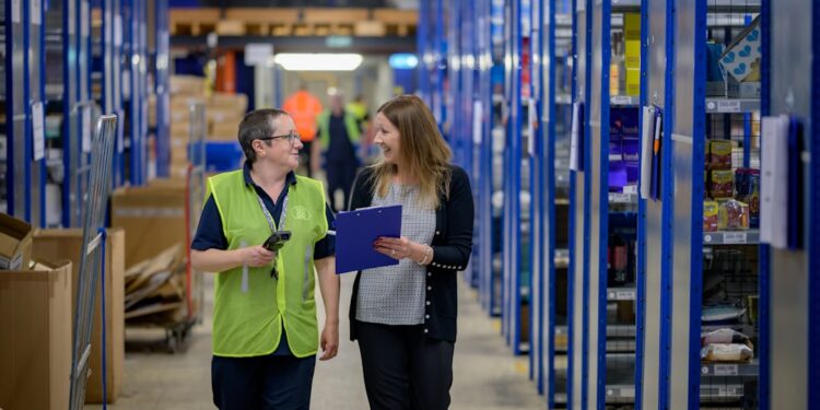 Two women walking through a warehouse and talking. One is holding a clipboard.