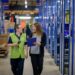 Two women walking through a warehouse and talking. One is holding a clipboard.
