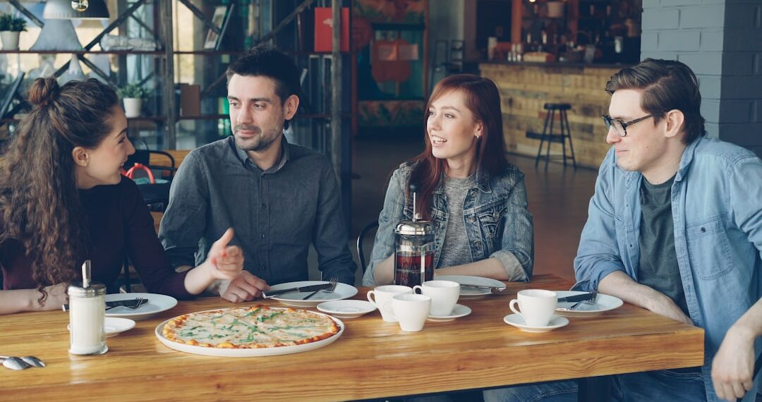 Cheerful young friends are talking and gesturing sharing news while sitting at table in modern cafe together. Big pizza, cups and plates, tables and chairs are visible.