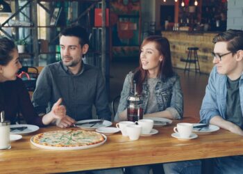 Cheerful young friends are talking and gesturing sharing news while sitting at table in modern cafe together. Big pizza, cups and plates, tables and chairs are visible.