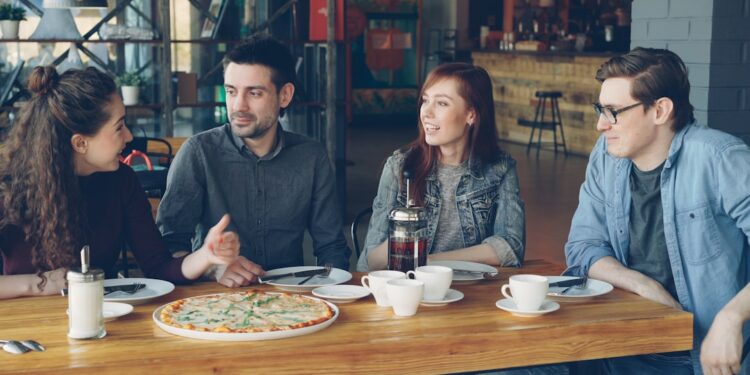 Cheerful young friends are talking and gesturing sharing news while sitting at table in modern cafe together. Big pizza, cups and plates, tables and chairs are visible.