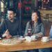 Cheerful young friends are talking and gesturing sharing news while sitting at table in modern cafe together. Big pizza, cups and plates, tables and chairs are visible.