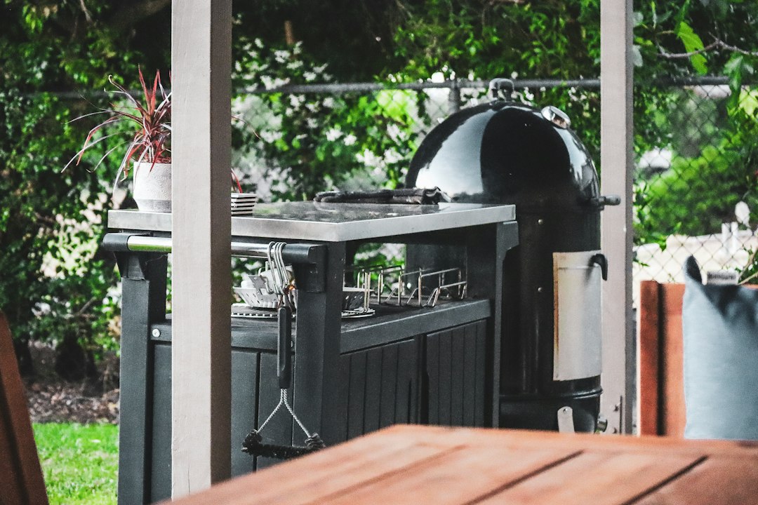 Zwarte rookoven naast een donkere buitenkast met rvs werkblad onder een pergola