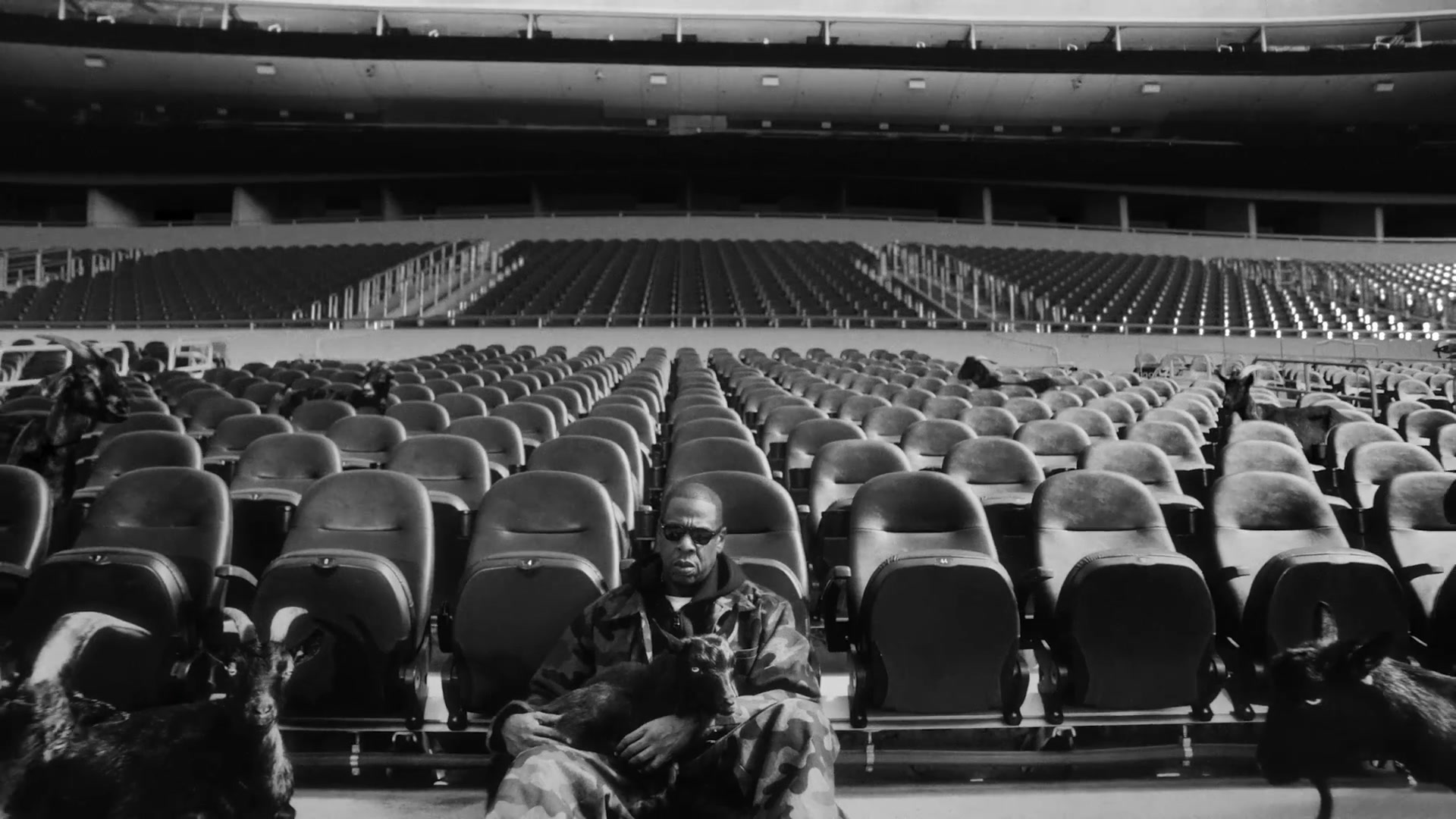 Jay-Z op de tribune van een leeg stadion tussen zwarte geiten in de aankondiging van de halftimeshow