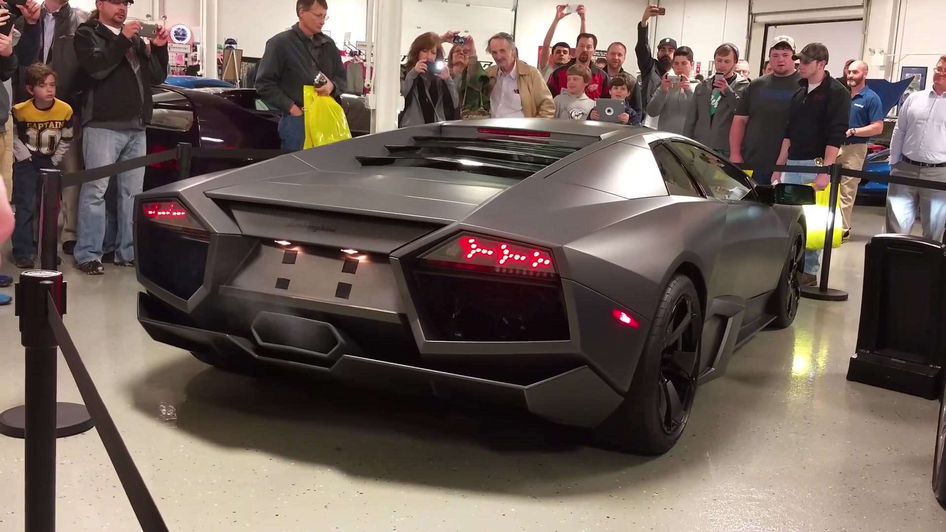 Een matgrijze Lamborghini Reventón van achteren, met de Y-vormige achterlichten aan, omringd door publiek